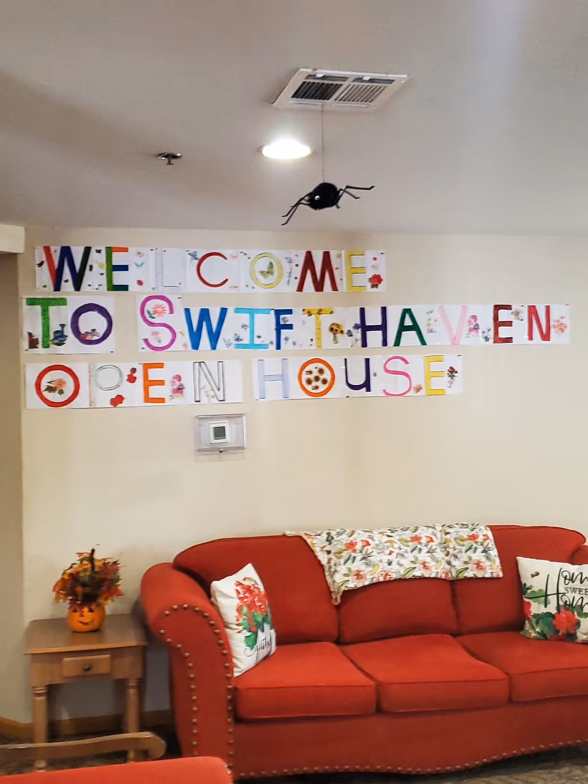 Interior view of a room with a red couch adorned with floral pillows and a floral throw blanket. Above the couch, colorful letters on paper spell out 'WELCOME TO SWIFTHAVEN OPEN HOUSE'. A small wooden side table next to the couch holds a pumpkin-shaped vase with autumn leaves. The ceiling has a light fixture and an air vent.