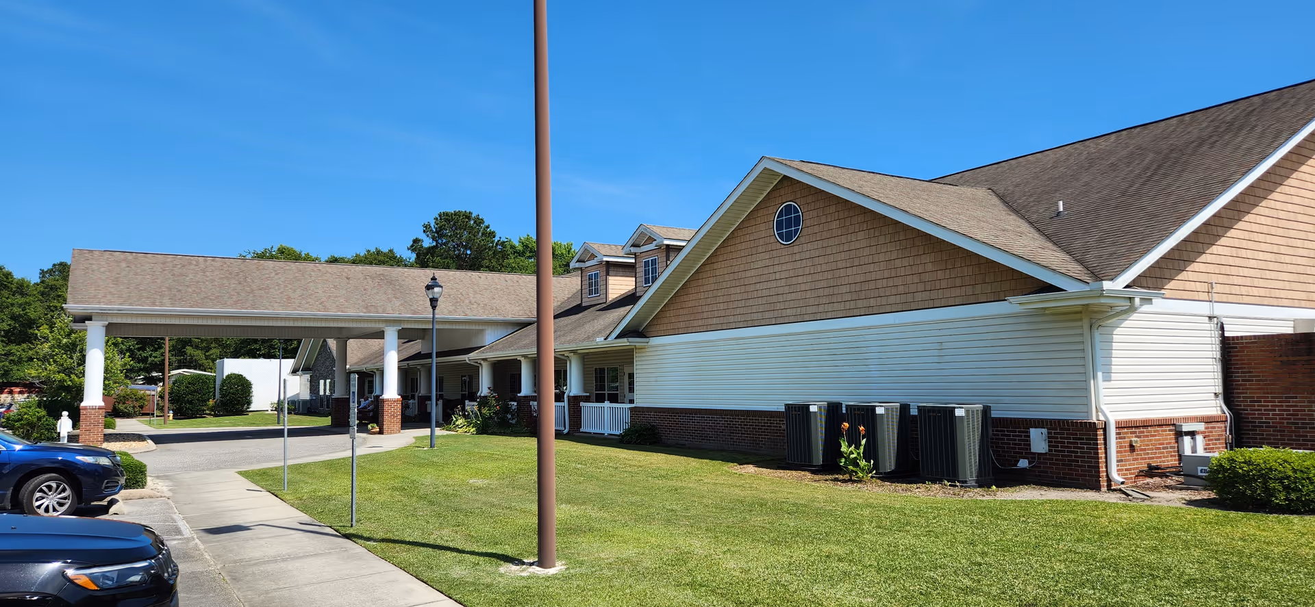 Exterior view of a single-story building with a covered entrance supported by white columns. The building has a combination of brick and siding with a shingled roof. There are several air conditioning units along the side of the building, a grassy lawn, and a few parked cars visible. The sky is clear and blue.