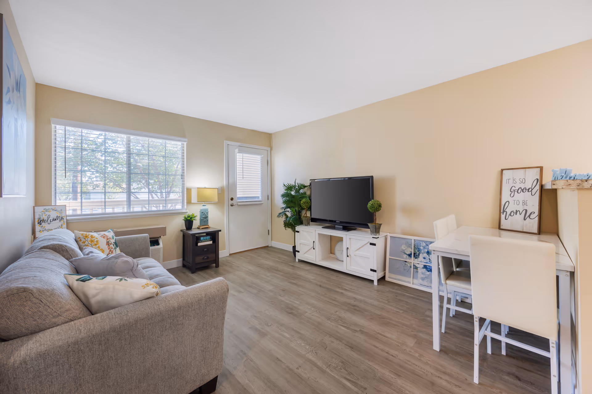 Well-lit living room with a gray sofa, TV on a white media console, a small dining table, and a window letting in natural light.
