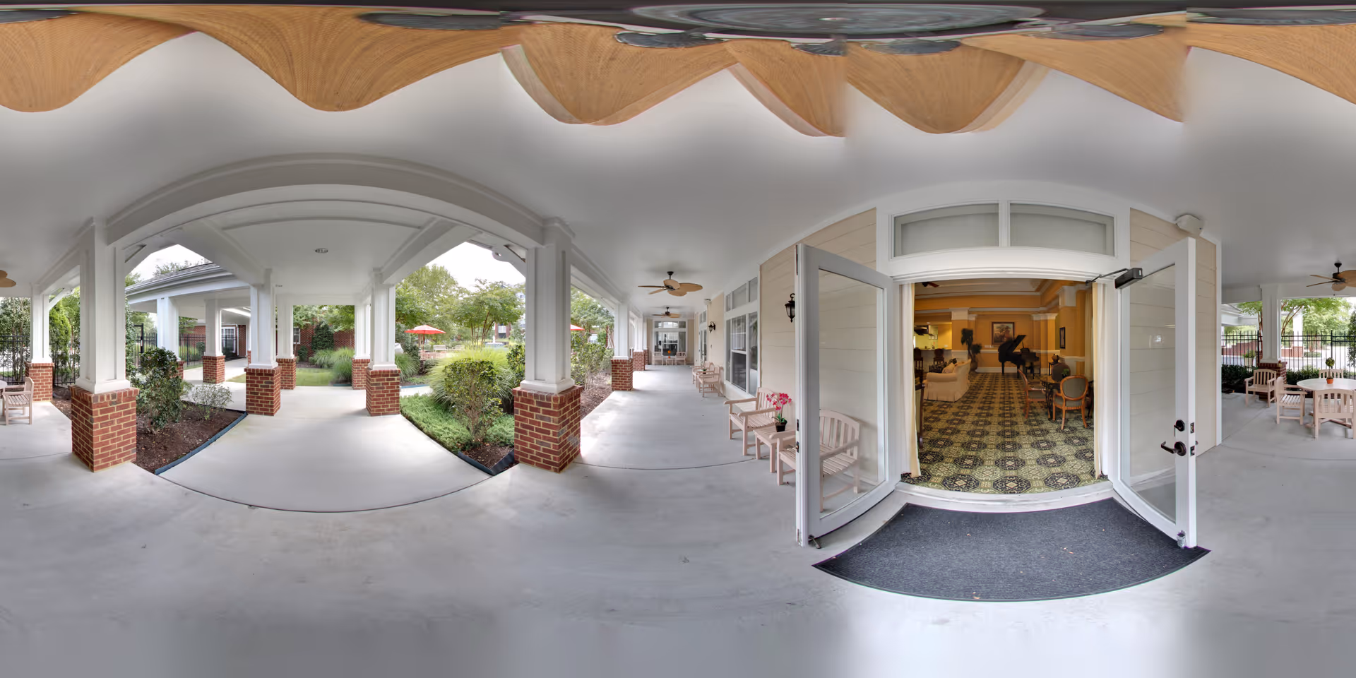 Covered outdoor walkway with white columns and brick bases leading to a garden area with greenery and red umbrellas. On the right side, double glass doors open into a room with patterned carpet, chairs, and a piano. Several benches and ceiling fans are visible along the walkway.