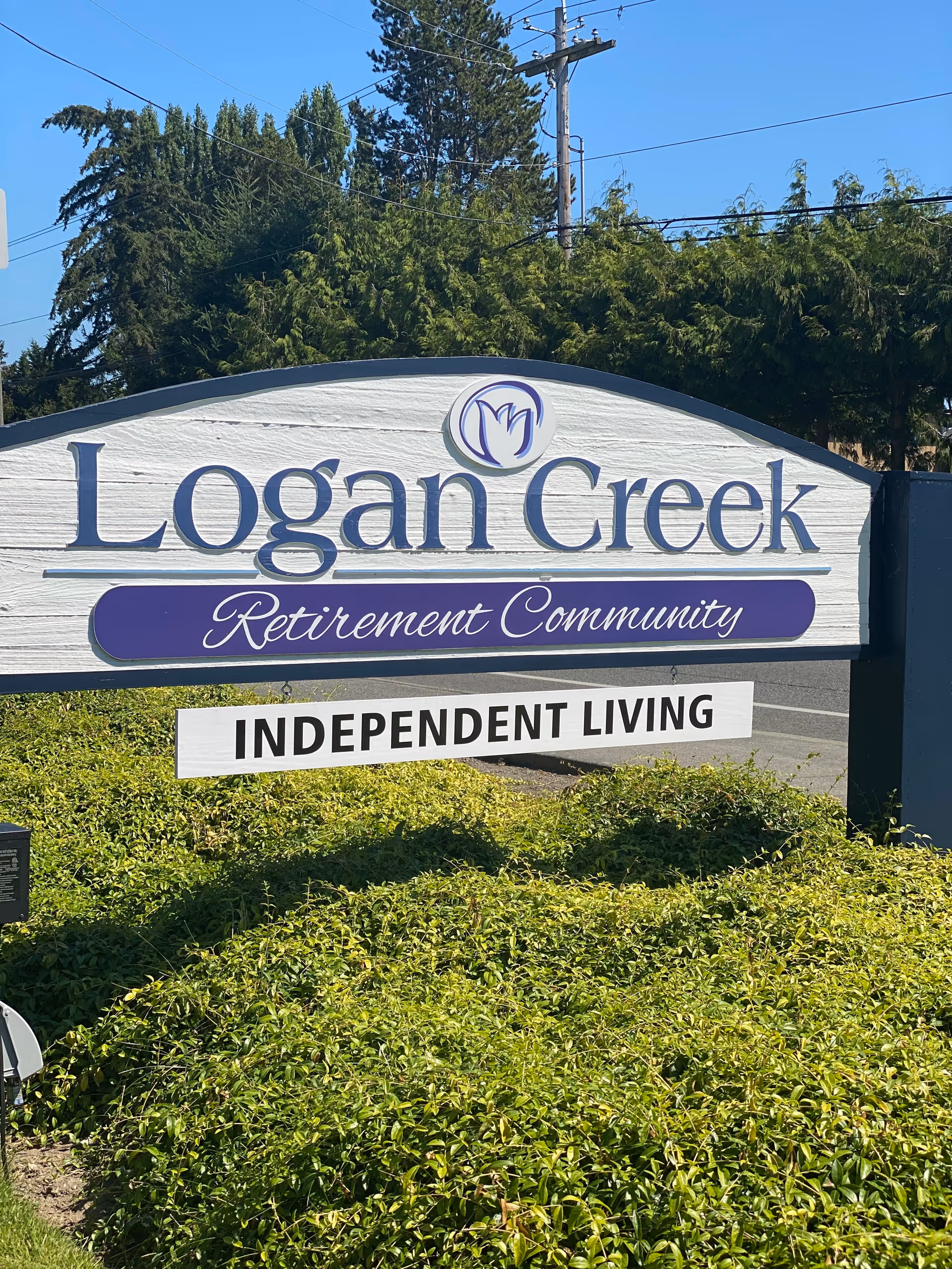 Outdoor sign for Logan Creek Retirement Community with the words Independent Living below it, surrounded by greenery and trees in the background under a clear blue sky.
