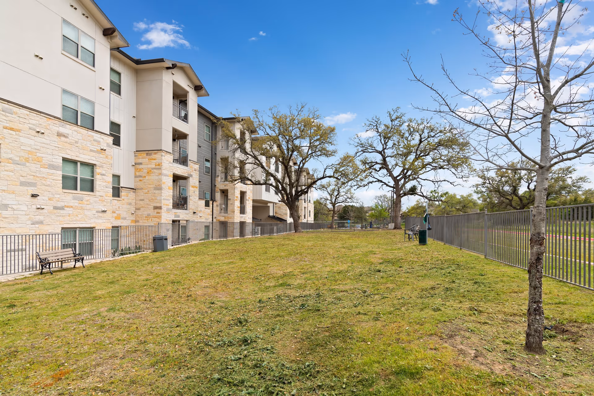 Outdoor grassy area next to a multi-story building with stone and beige siding. There are several trees, a metal fence, benches, and a trash can under a blue sky with some clouds.