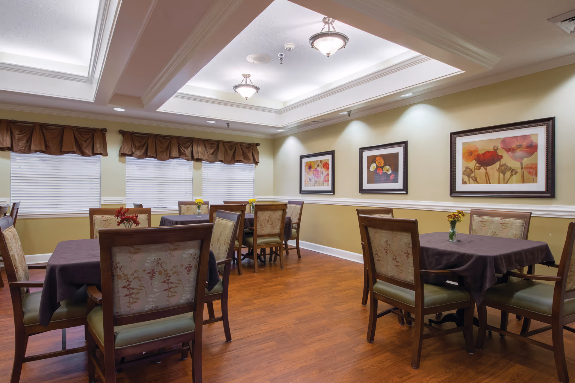 A dining room with several tables covered in dark tablecloths, each surrounded by wooden chairs with floral upholstery. The room has a wooden floor, light green walls with white trim, three framed floral paintings on the wall, and three windows with brown valances. The ceiling features recessed lighting and two hanging light fixtures.