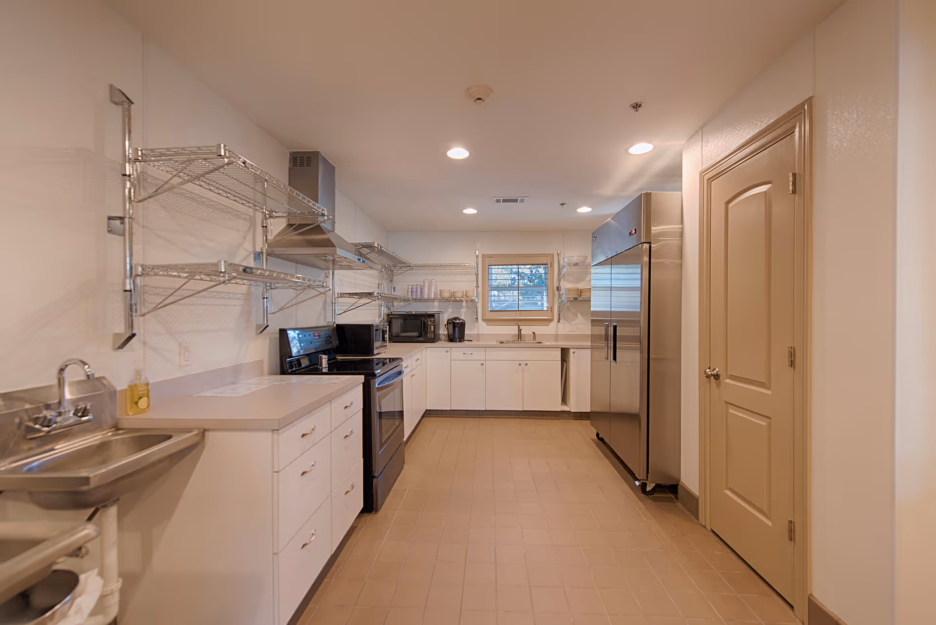 A clean, well-lit kitchen area with white cabinets, stainless steel appliances including a large refrigerator and stove, a small window above the sink, and metal wire shelving on the walls.