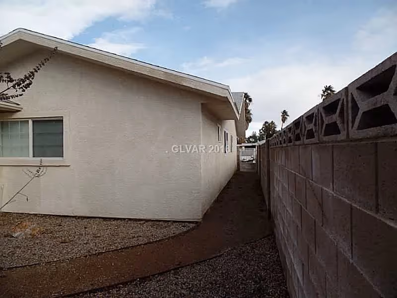 Narrow outdoor pathway alongside a beige building with a window on the left and a concrete block wall on the right, under a partly cloudy sky.
