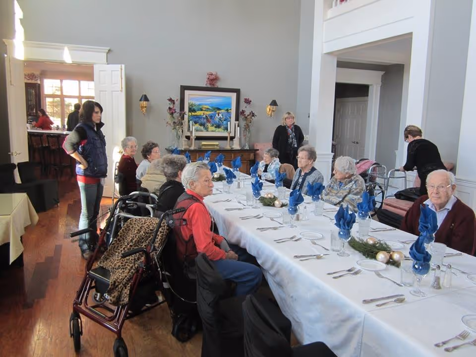 A group of elderly people seated around a long dining table set with white tablecloth, blue folded napkins, plates, glasses, and silverware in a well-lit room. Two women stand nearby, and a walker is visible next to one of the seated individuals. The room has wooden floors, light-colored walls, a painting, and decorative wall sconces.