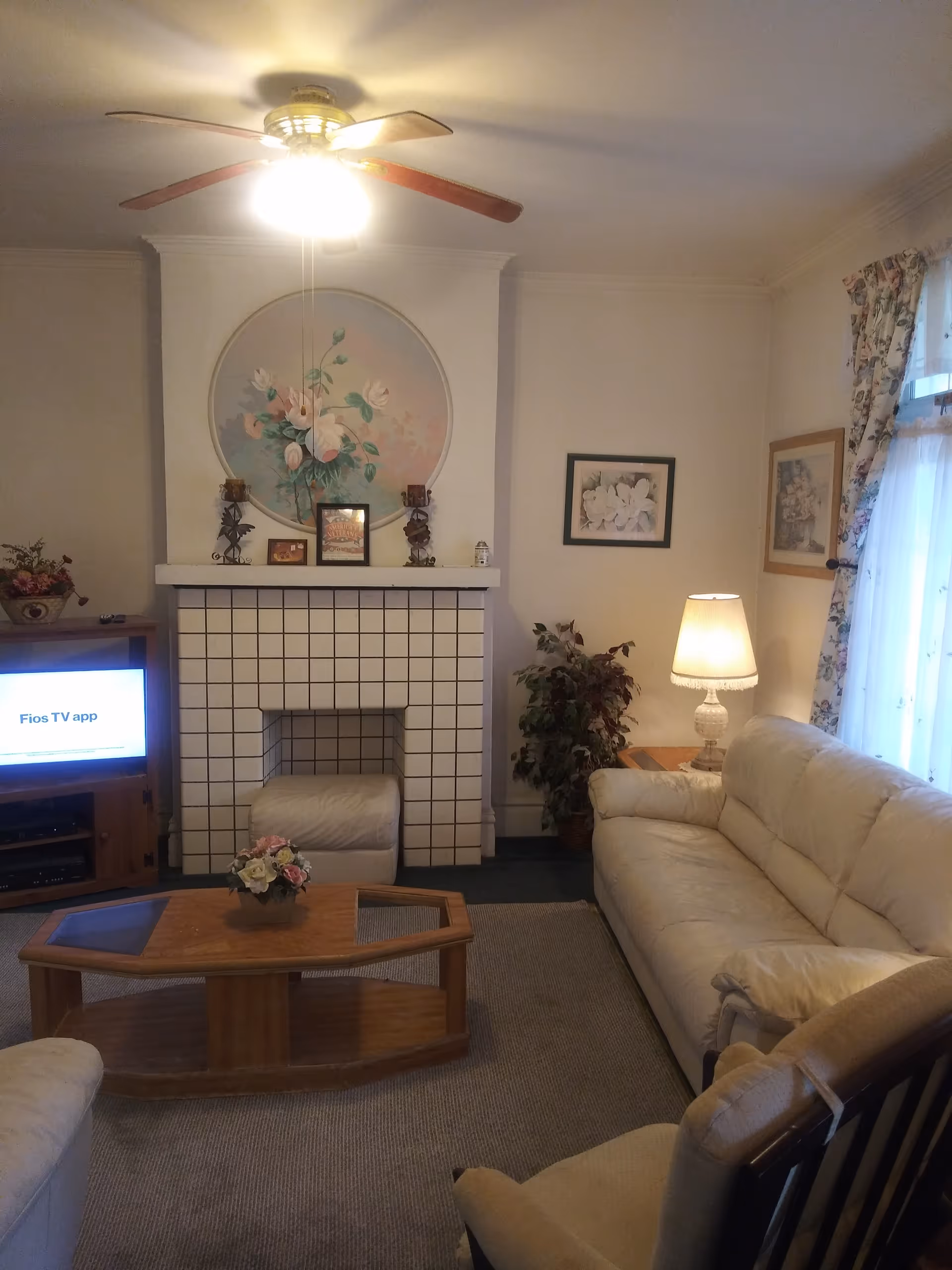Living room with cream sofas, wooden coffee table, tiled fireplace topped by a floral mural, TV, lamp, and ceiling fan.