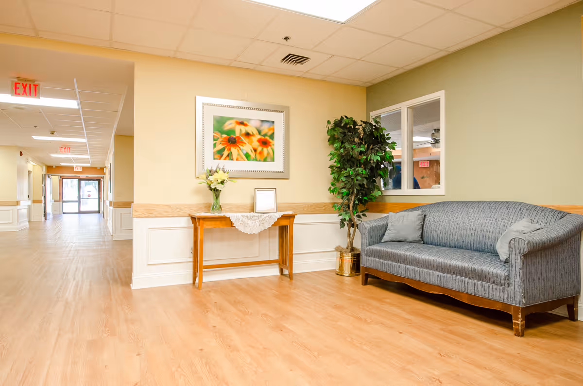 A bright senior facility interior with a blue sofa, a small console table holding flowers and framed artwork, and a hallway leading to an exit.