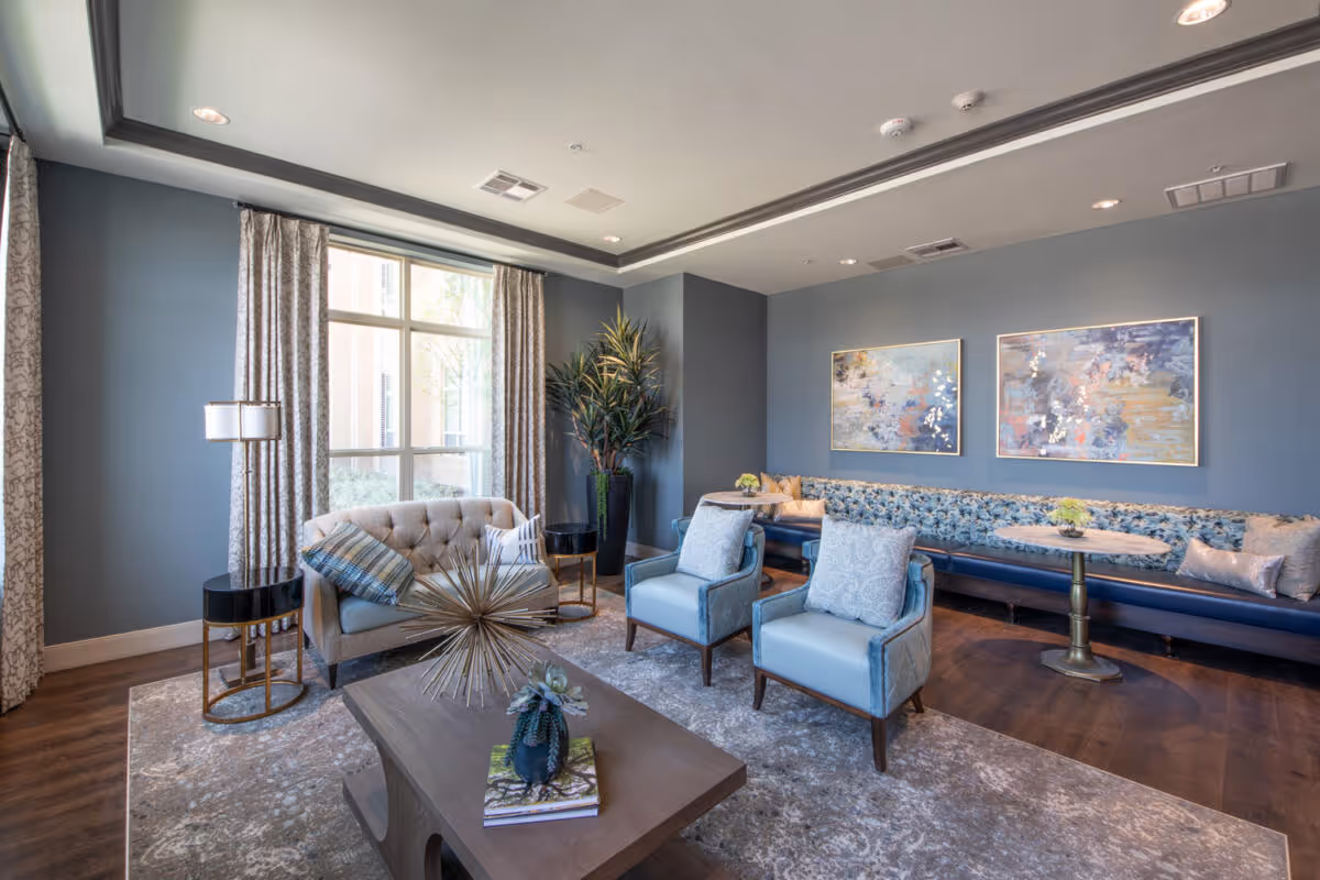 A modern and cozy living room area with blue walls and a large window with patterned curtains. The room features a tufted beige loveseat with pillows, two light blue armchairs with patterned cushions, a long blue cushioned bench along the wall with small round tables, and decorative abstract paintings hanging above the bench. A wooden coffee table with decorative items sits on a patterned rug over wooden flooring.