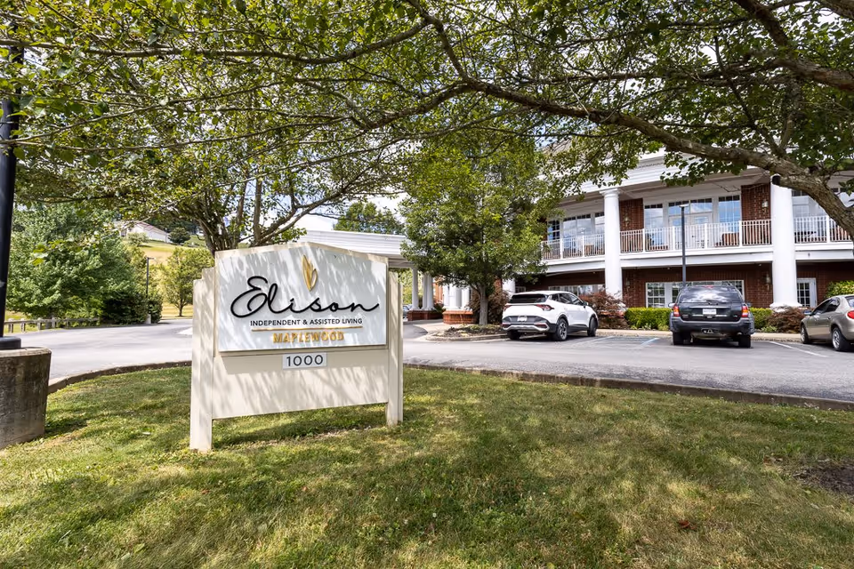 Outdoor view of Elison Independent & Assisted Living of Maplewood facility sign with the building and parked cars in the background, surrounded by trees and grass.