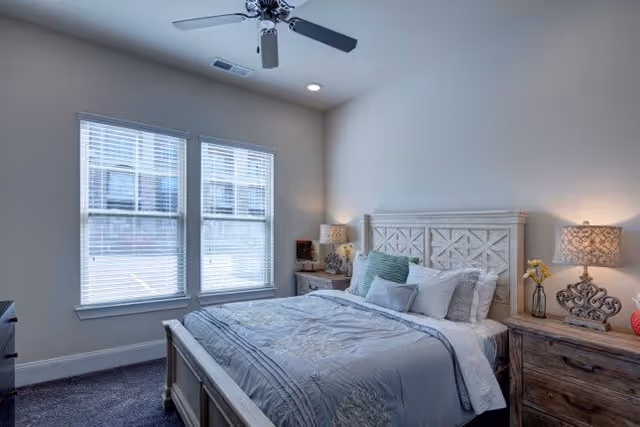 A bright bedroom with a large bed featuring a decorative headboard, multiple pillows, and a gray and white comforter. There are two wooden nightstands on either side of the bed, each with a lamp and decorative items. Two large windows with blinds allow natural light into the room, and a ceiling fan is mounted above.