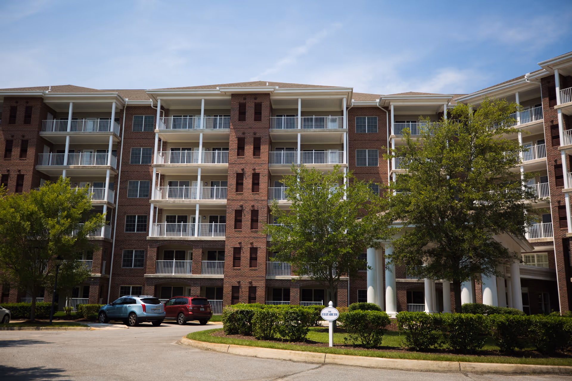 Front view of a multi-story brick senior living building with balconies, trees, parked cars, and an entrance sign.