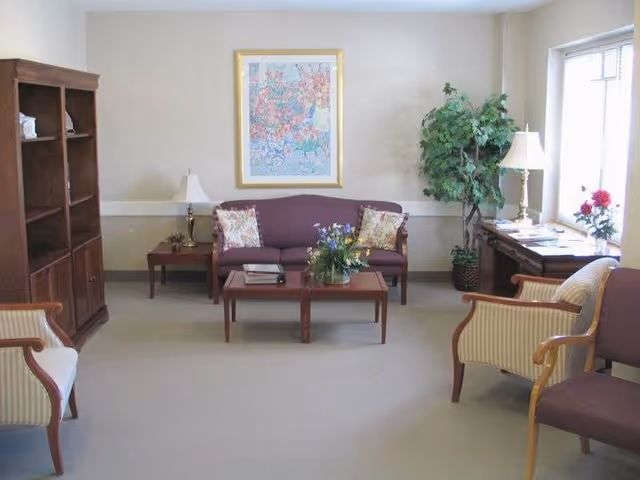 A cozy living room area with a purple sofa, two striped armchairs, a wooden coffee table with a flower arrangement, a wooden bookshelf, two side tables with lamps, a large framed floral artwork on the wall, and a potted plant near a window with natural light.