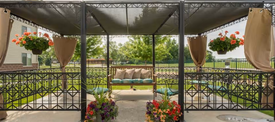 Covered outdoor patio area with a metal frame and roof, featuring a cushioned bench with pillows in the center, hanging flower baskets with orange flowers, potted colorful flowers on the ground, and beige curtains tied to the metal posts. Green grass and trees are visible in the background.