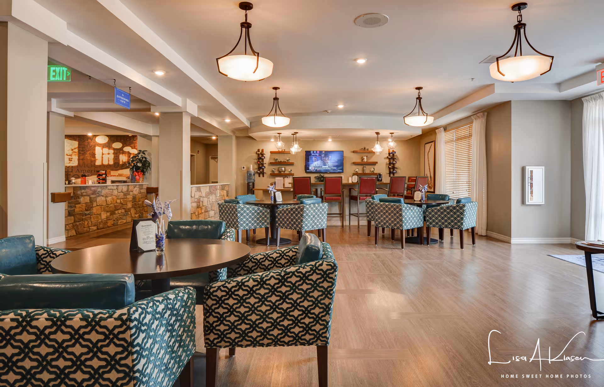 A spacious senior living common area with multiple round tables surrounded by patterned and solid teal chairs. The room features warm lighting from ceiling fixtures, wood flooring, and large windows with blinds and curtains allowing natural light. In the background, there is a counter area with stone facing and a mounted TV on the wall above a bar with red chairs. The space is decorated with small plants and shelves holding decorative items.