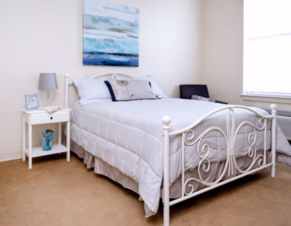 A neatly made bed with a white metal frame and light gray bedding in a bedroom. There is a white nightstand with a gray lamp and a small framed picture on it. A blue decorative item is on the lower shelf of the nightstand. A piece of abstract artwork with blue tones hangs on the wall above the bed. A window with blinds is on the right side, and a dark chair is positioned in the corner near the window.