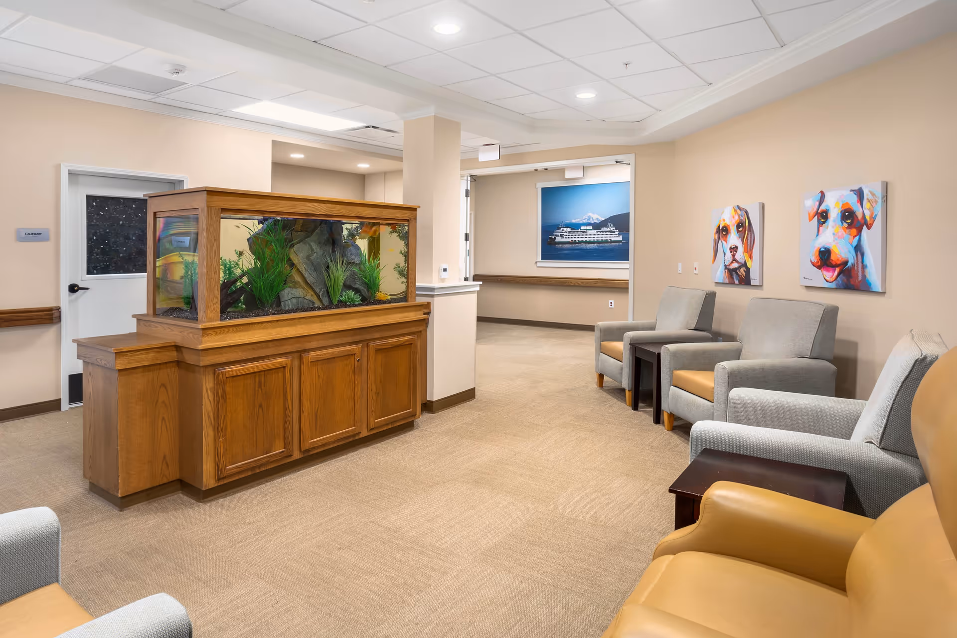 A senior living facility seating area with an aquarium on a wooden cabinet, several armchairs, and colorful dog artwork on the wall.