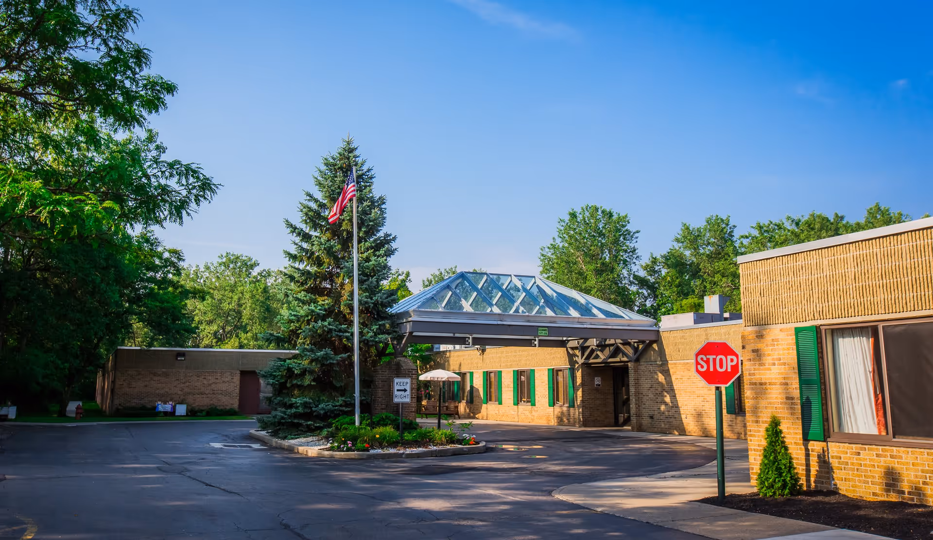 Exterior view of Seneca Health Care Center showing a brick building with green window shutters, a glass canopy entrance, an American flag on a flagpole, a stop sign, and surrounding trees under a clear blue sky.