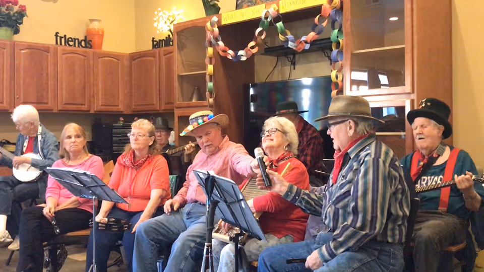A group of elderly people sitting in a living room area, some holding musical instruments such as a banjo and tambourine, and one person holding a microphone. The room has wooden cabinets, a TV, and colorful paper chains hanging as decorations.
