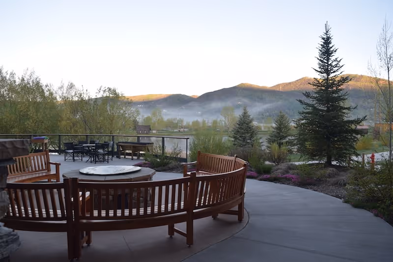 Outdoor patio area with curved wooden benches surrounding a circular stone fire pit, additional tables and chairs in the background, with trees, shrubs, and mountains visible in the distance under a clear sky.