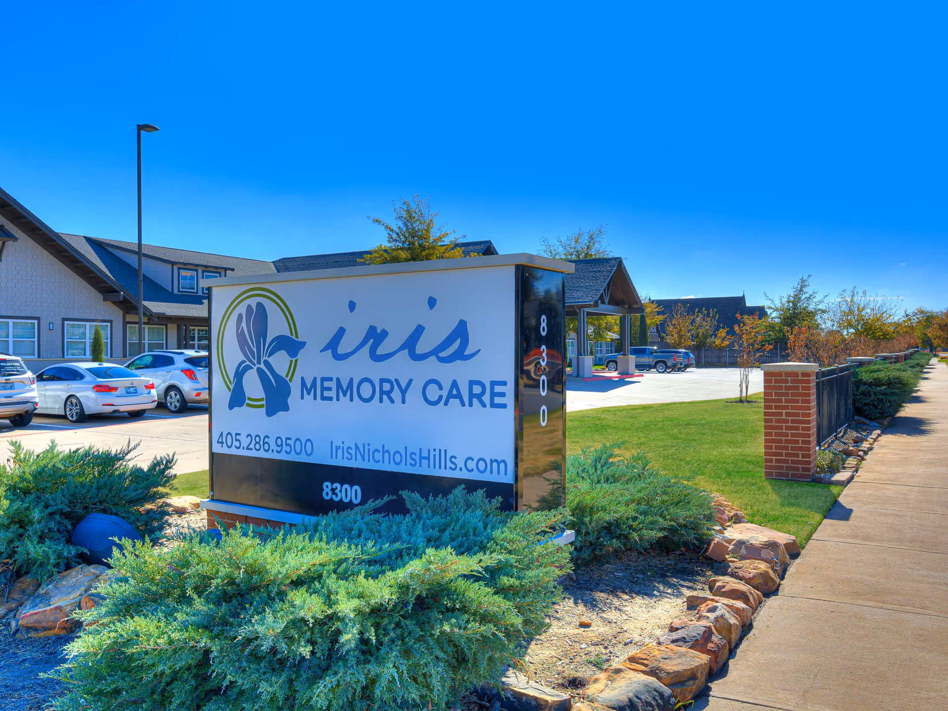 Outdoor view of Iris Memory Care of Nichols Hills facility sign with a landscaped area featuring bushes and rocks in the foreground, a parking lot with several cars, and the building in the background under a clear blue sky.