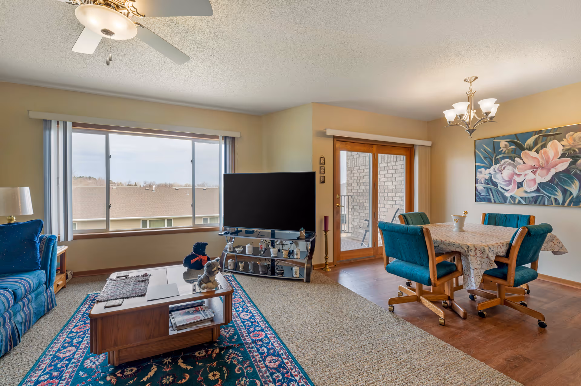 A cozy senior living room and dining area with a large window and glass doors leading to a balcony. The living room has a blue striped sofa, a wooden coffee table with stuffed animals, a flat-screen TV on a stand, and a ceiling fan with a light. The dining area features a wooden table with a floral tablecloth and four teal cushioned chairs on wheels, a chandelier overhead, and a large floral painting on the wall.