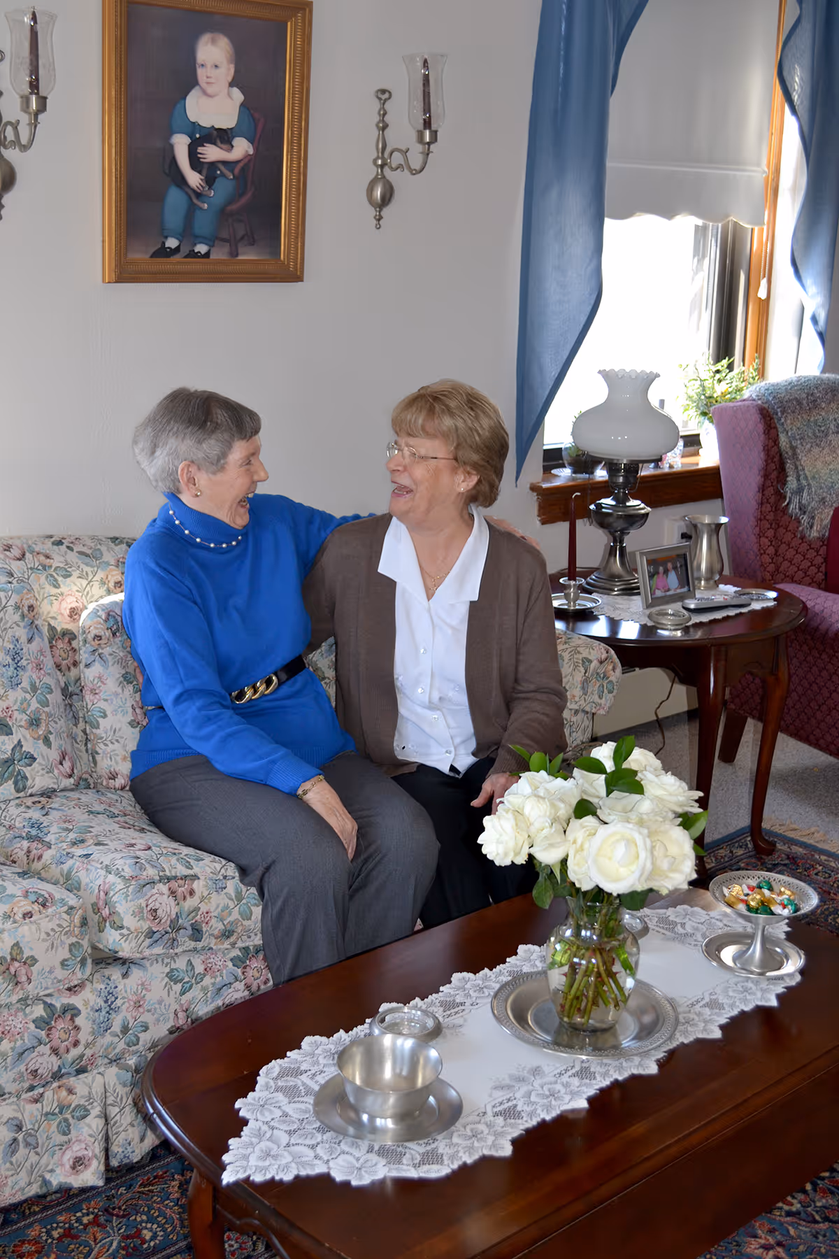 Two elderly women sitting on a floral patterned couch in a cozy living room, smiling and embracing each other. The room features a wooden coffee table with a lace runner, a vase of white flowers, silver cups, and a bowl of candy. Behind them is a side table with a lamp, framed photos, and a window with blue curtains. A portrait of a child holding a cat hangs on the wall above the couch.