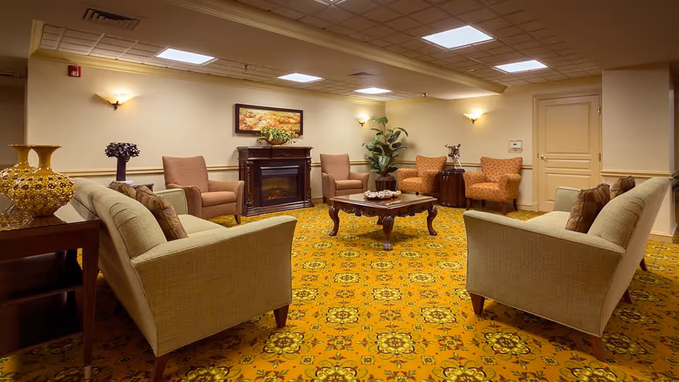 A cozy senior living community lounge with beige sofas and armchairs arranged around a wooden coffee table. The room features a patterned yellow carpet, a fireplace with a floral painting above it, wall sconces providing warm lighting, and decorative plants and vases.