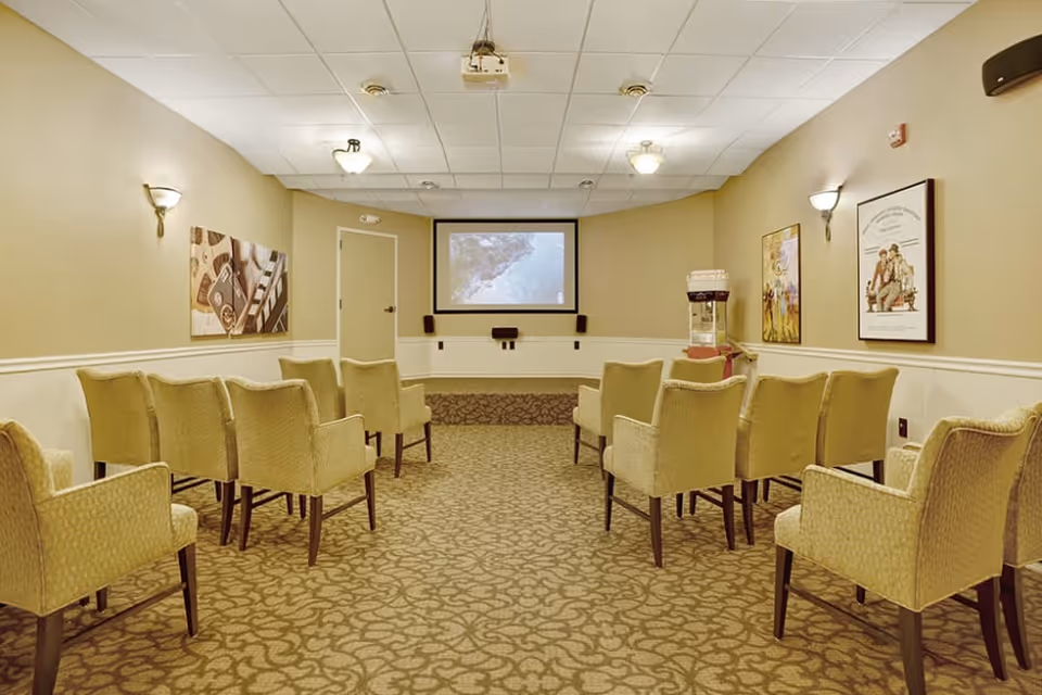 A small theater room with two rows of beige upholstered chairs facing a large screen on the front wall. The room has beige walls with white wainscoting, carpeted floor with a patterned design, wall sconces for lighting, and framed posters on the walls. A popcorn machine is visible in the corner near the screen.