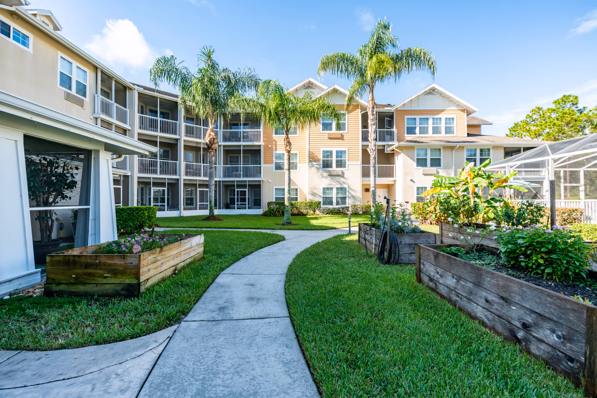 Courtyard with raised garden beds, a curved walkway, palm trees, and a three-story residential building under a blue sky.