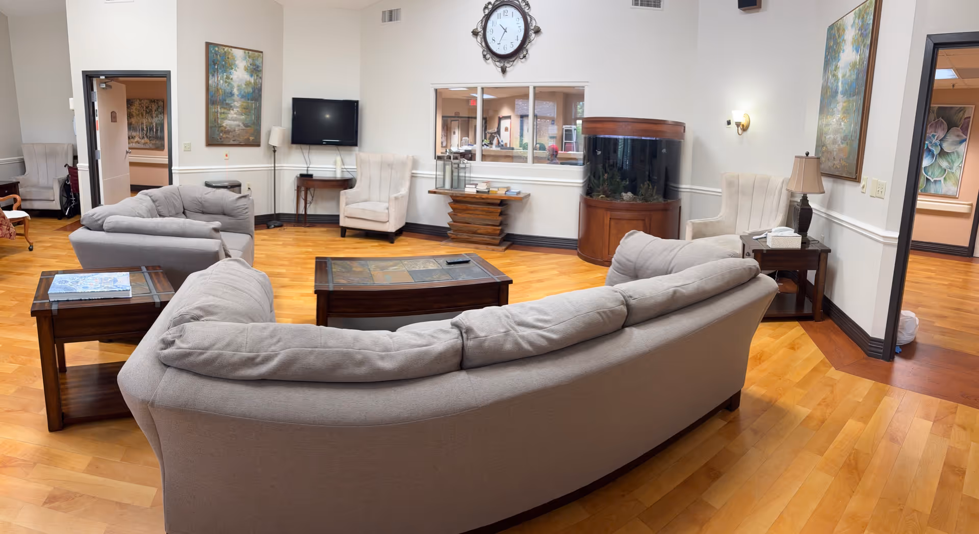 A cozy senior living common area with light gray sofas and armchairs arranged around a wooden coffee table. The room features hardwood floors, a wall-mounted TV, a large round fish tank, a clock above the tank, and framed artwork on the walls. There are windows and doorways leading to other rooms.