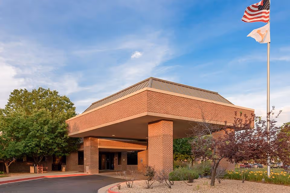 Exterior view of a brick building with a covered entrance supported by pillars, surrounded by trees and landscaping. An American flag and another flag are flying on a flagpole to the right under a partly cloudy blue sky.