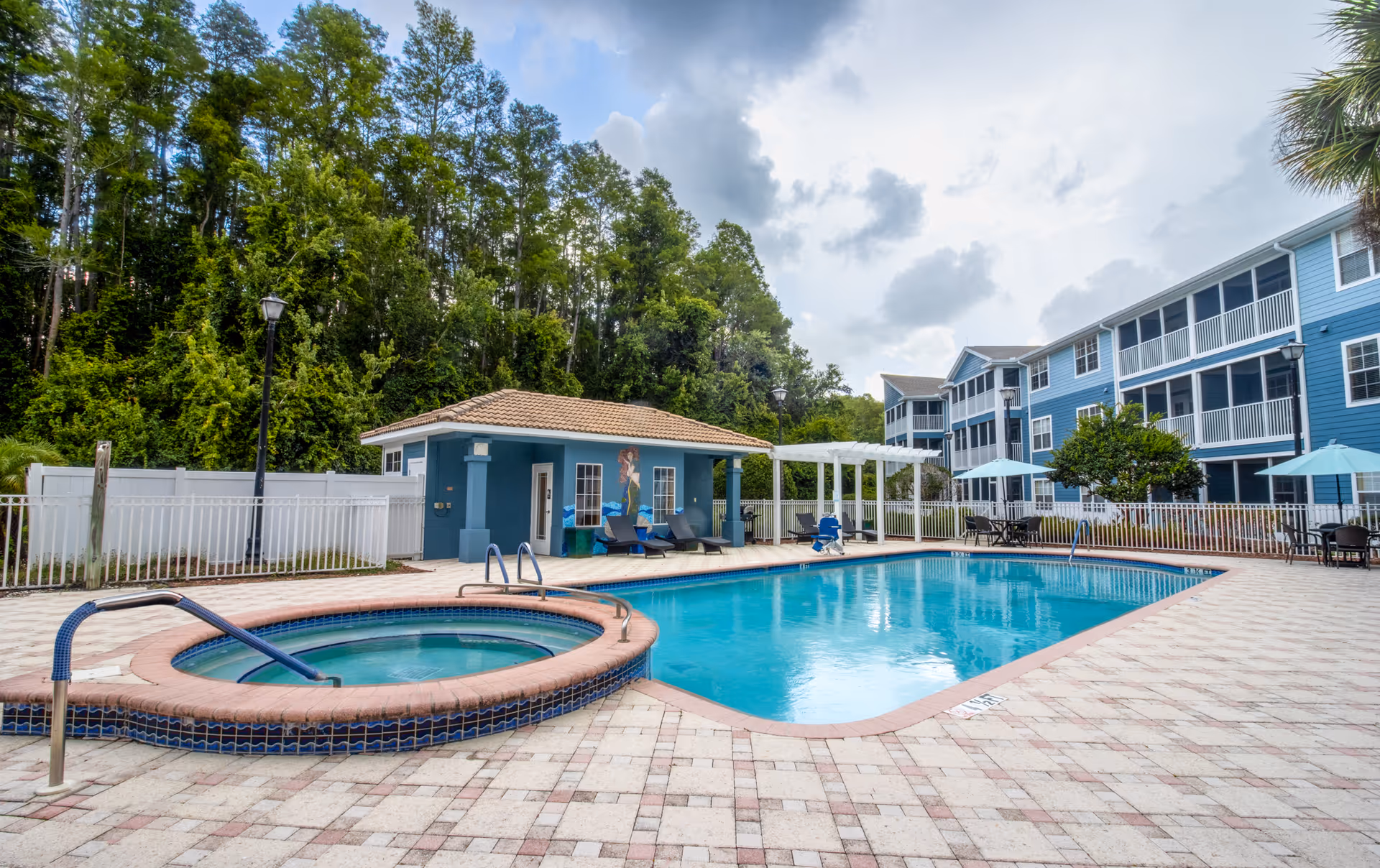 Outdoor swimming pool and hot tub area at Addington Place of East Lake, surrounded by a tiled deck with lounge chairs, tables with umbrellas, and a small blue building with a tiled roof. Trees and a multi-story blue residential building are visible in the background under a partly cloudy sky.