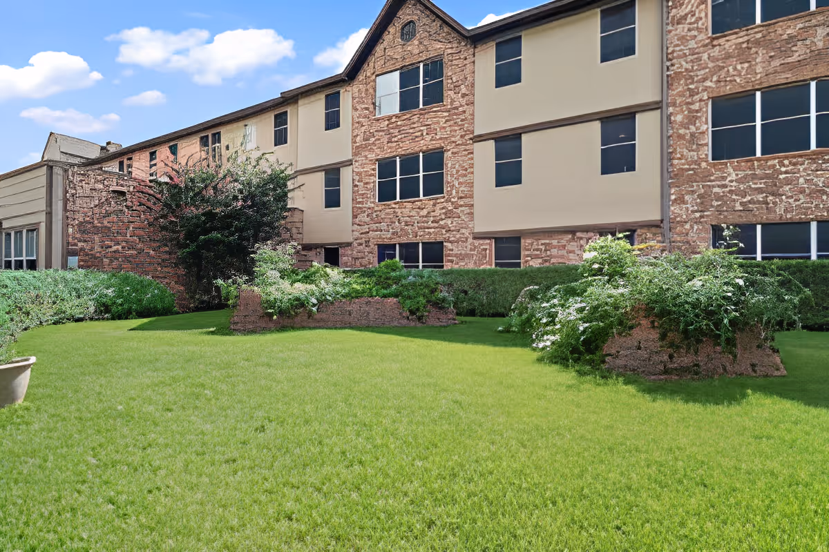 Outdoor view of Morada Meadow Lakes facility showing a well-maintained green lawn with bushes and plants in front of a multi-story building with stone and beige exterior walls under a blue sky with some clouds.