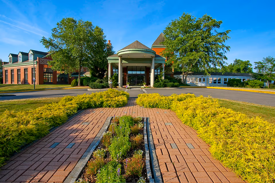 Front exterior view of a senior living facility with a brick walkway leading to the main entrance, flanked by yellow shrubs and green trees under a clear blue sky.