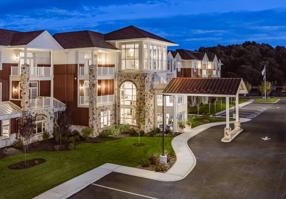 Exterior view of Fieldstone at Chester Springs senior living facility at dusk, showing a large building with stone and wood paneling, multiple balconies, well-lit windows, a covered entrance, landscaped greenery, and a parking area.