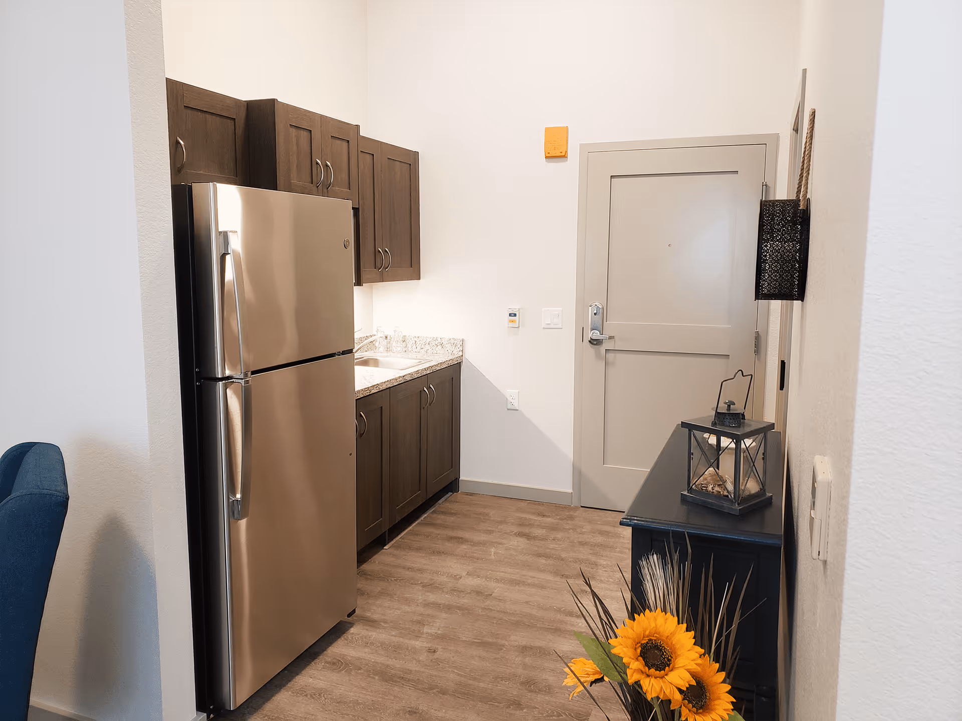 A small kitchen area with a stainless steel refrigerator, dark wooden cabinets, and a granite countertop with a sink. To the right is a closed door, a black console table with a decorative lantern, and a vase with sunflowers and dried plants. The floor is wood, and the walls are white.