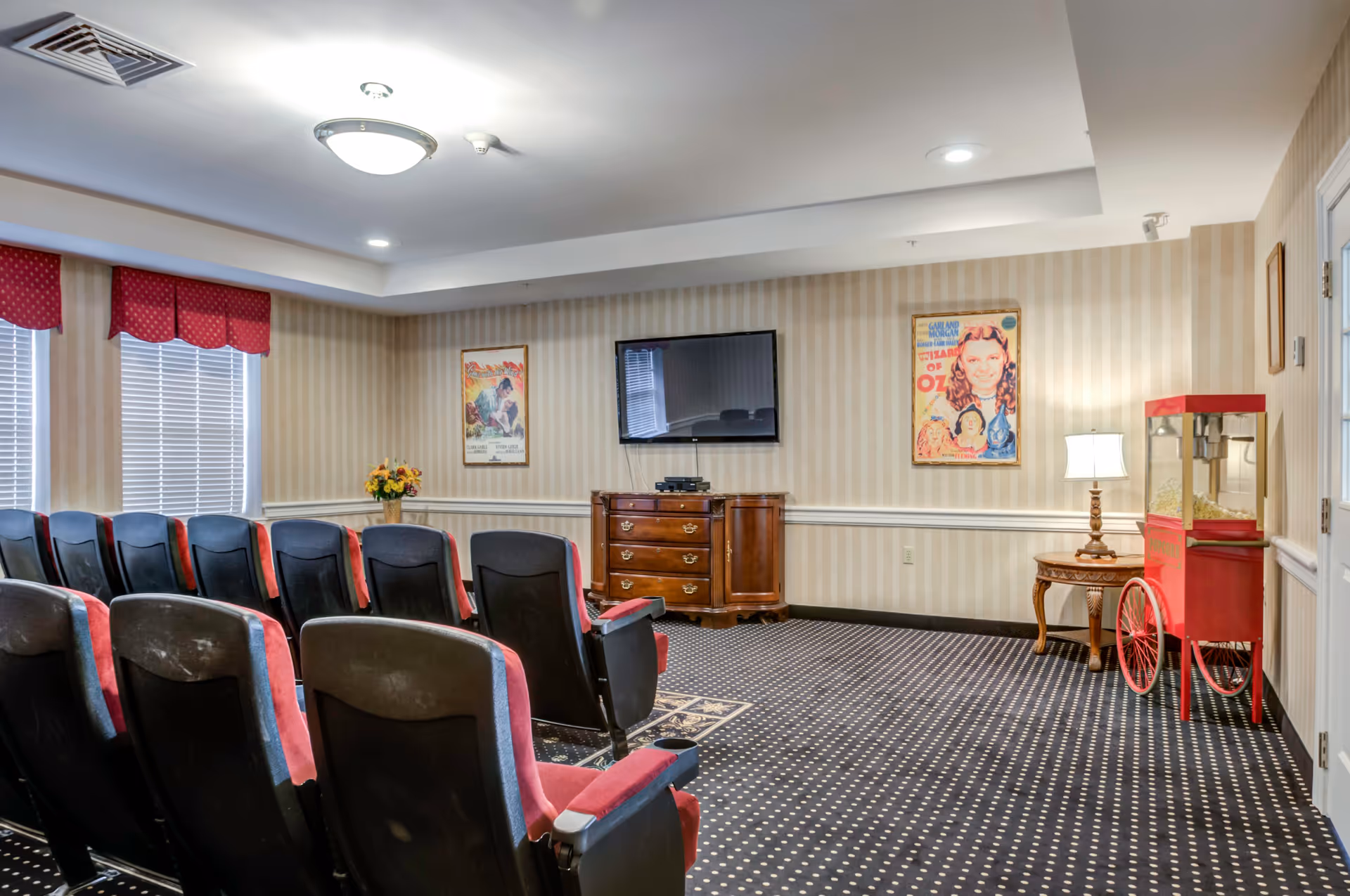 A small movie theater room with rows of black and red cushioned seats facing a flat-screen TV mounted on a striped wallpapered wall. There are two vintage movie posters on the wall, a wooden cabinet under the TV, a red popcorn machine on wheels, a small wooden side table with a lamp, and windows with red valances.