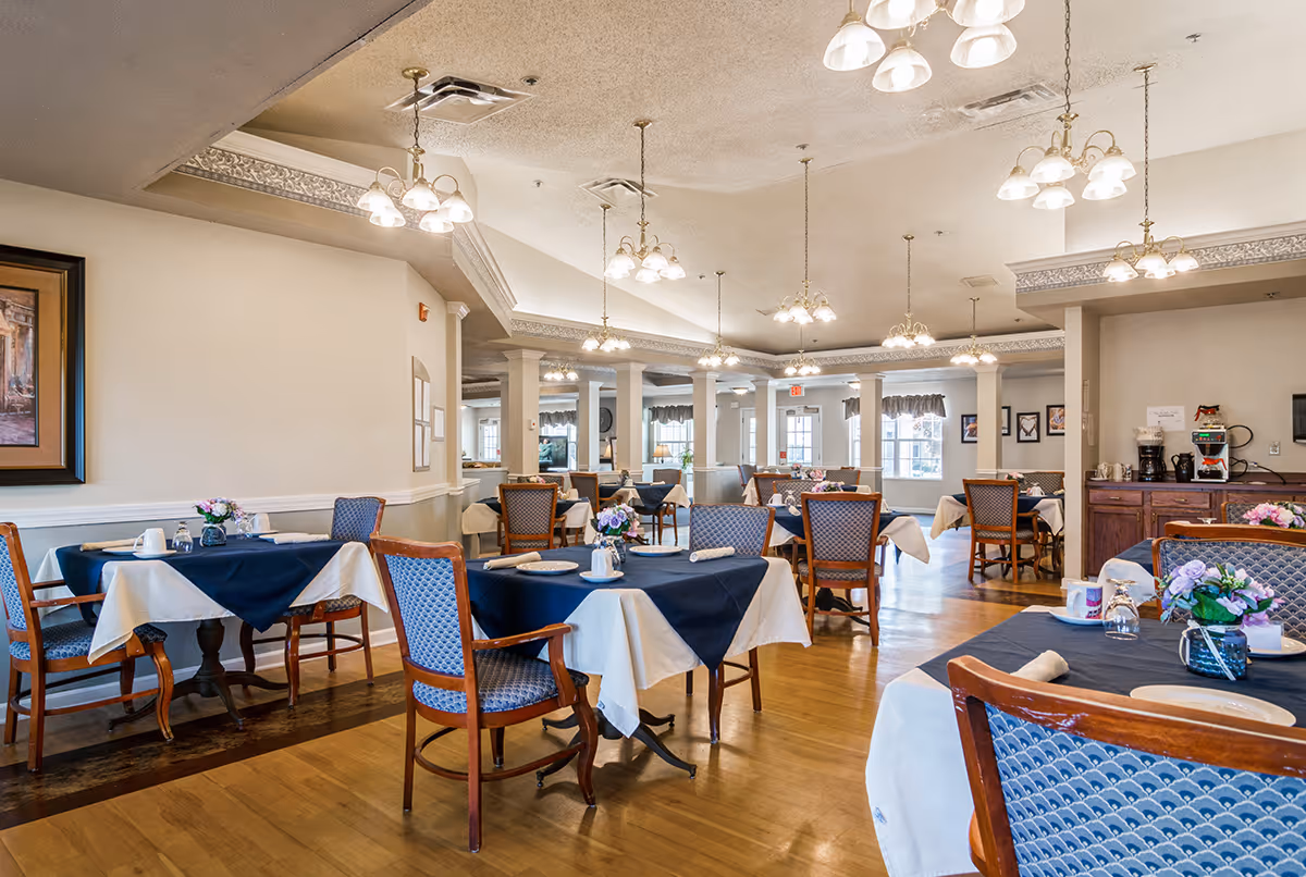 A spacious dining room with multiple tables covered in white and navy blue tablecloths, each set with plates, cups, and napkins. The room has wooden chairs with blue patterned upholstery, hardwood floors, and several hanging light fixtures. There are windows with valances letting in natural light and framed artwork on the walls.