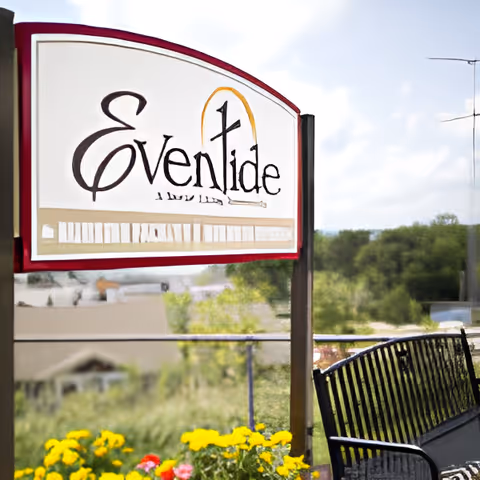 Outdoor view of a sign for EvenTide, an Eventide Community, with a black metal bench nearby and yellow flowers in the foreground. Trees and buildings are visible in the background under a partly cloudy sky.