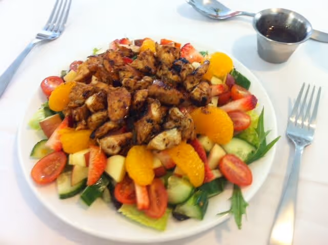 A plate of salad with grilled chicken pieces on top, surrounded by sliced cucumbers, cherry tomatoes, mandarin orange segments, strawberries, and apple chunks. A small metal container of dressing is placed next to the plate on a white tablecloth, with a fork on each side of the plate.