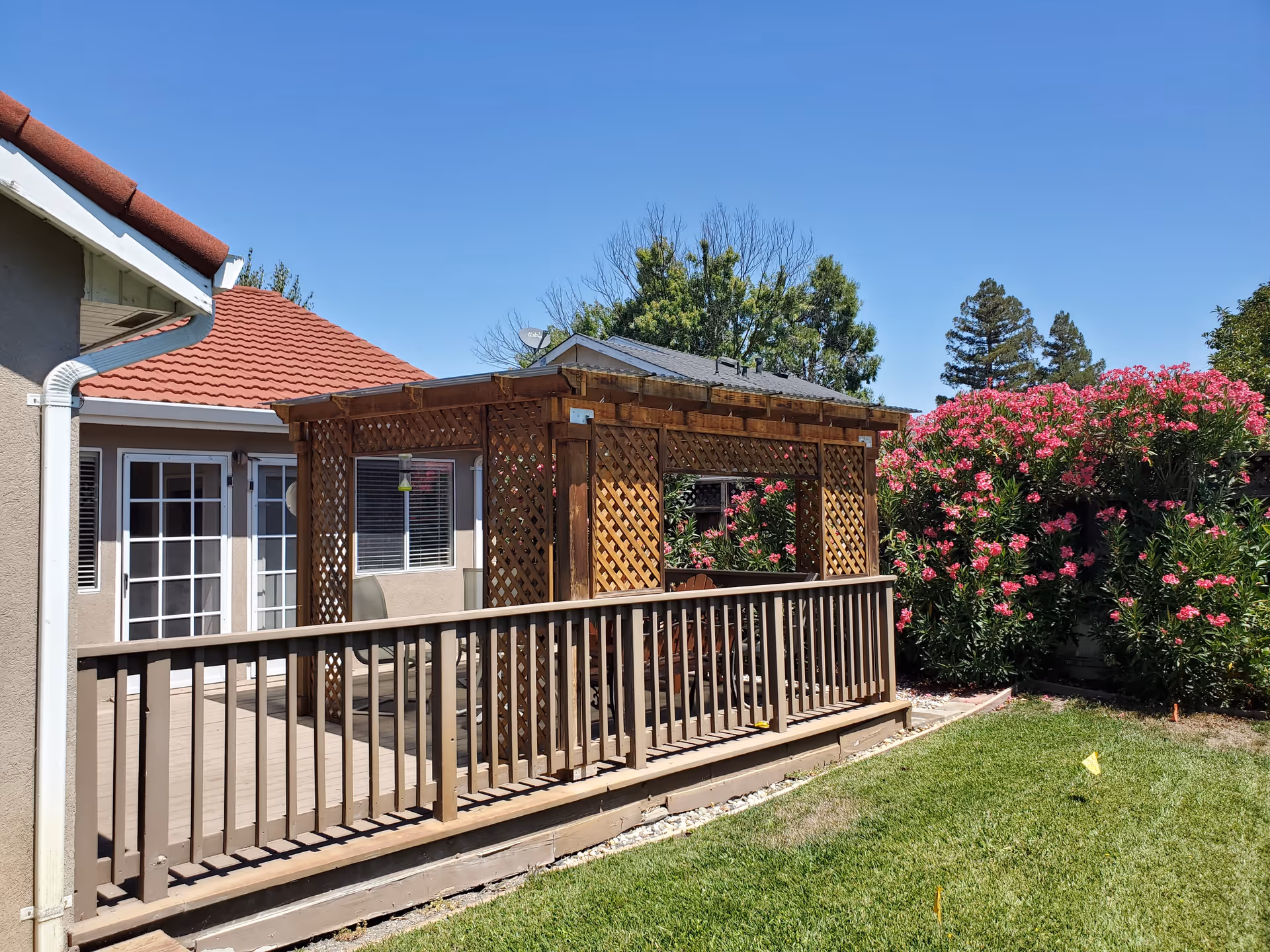 Outdoor patio area with a wooden pergola and railing attached to a building with red tiled roof. There are pink flowering bushes and green grass surrounding the patio under a clear blue sky.