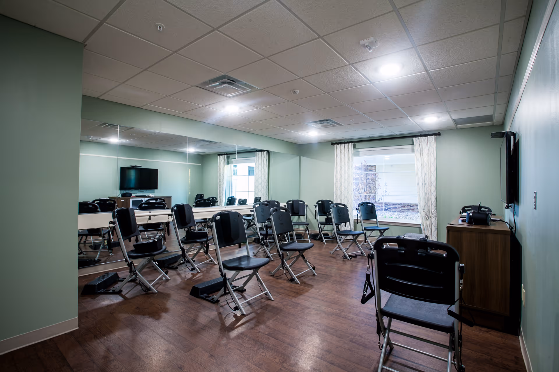 A room with multiple black folding chairs arranged in rows facing a wall-mounted television. The room has light green walls, a large mirror on one side, a window with patterned curtains, and wooden flooring. There is a small wooden cabinet beneath the television.