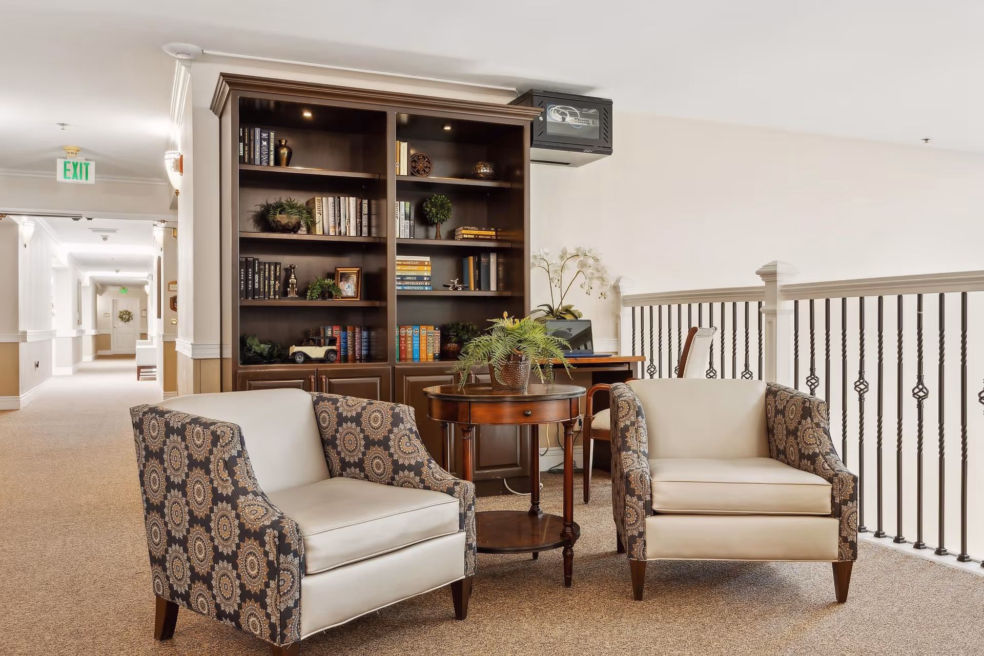 A cozy seating area in a senior living facility with two patterned armchairs and a round wooden table with a potted plant. Behind the seating area is a dark wooden bookshelf filled with books and decorative items. A desk with a chair and a computer is positioned next to the bookshelf. The area overlooks a railing with decorative iron balusters, and a hallway extends into the background with an exit sign visible.