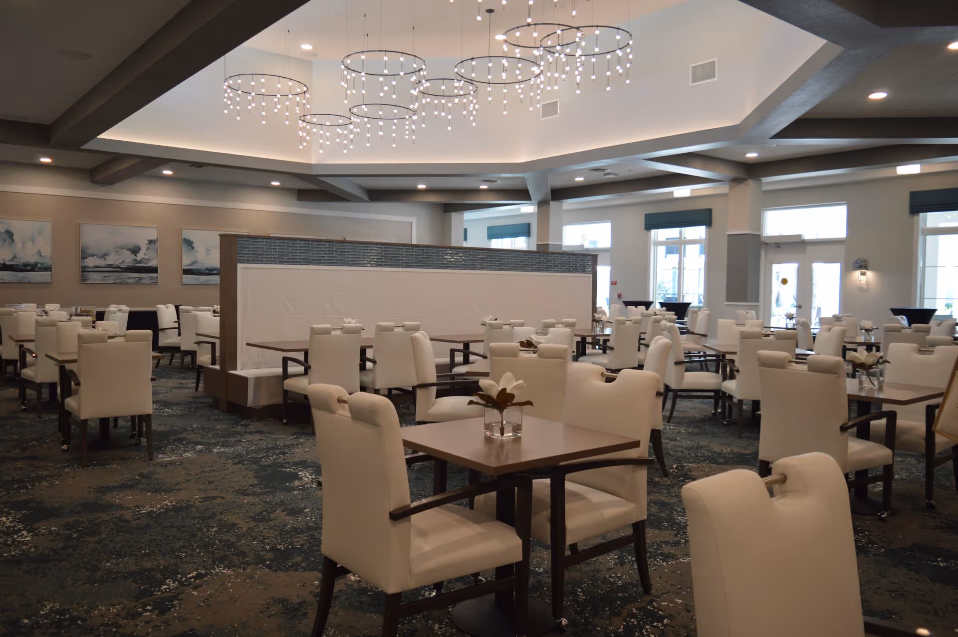 A spacious and well-lit dining room with multiple square wooden tables surrounded by white cushioned chairs. The room features modern circular chandeliers hanging from a high ceiling with recessed lighting. Large windows allow natural light to enter, and there are decorative paintings on the walls. The carpet has a patterned design in shades of blue and beige.