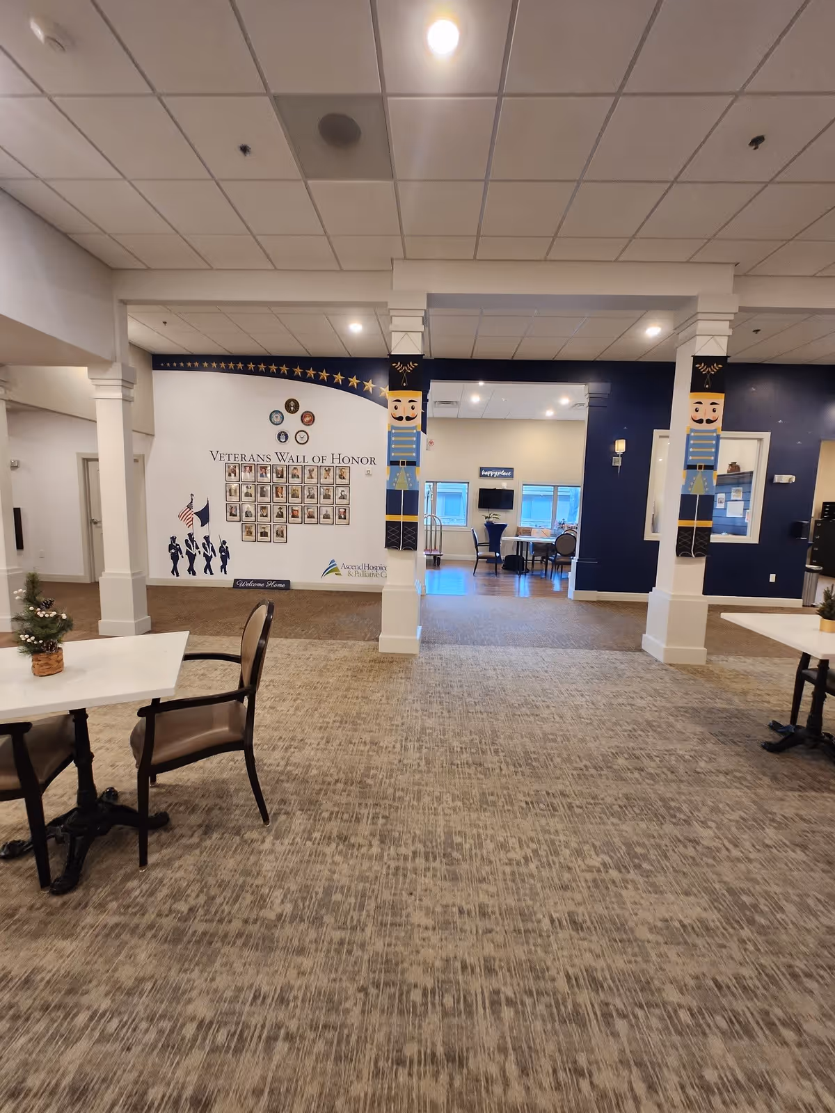 Interior view of a senior living facility featuring a Veterans Wall of Honor with photos and flags on the left wall. The space has carpeted flooring, white pillars decorated with nutcracker figures, and tables with chairs. In the background, there is a seating area with a television mounted on the wall.