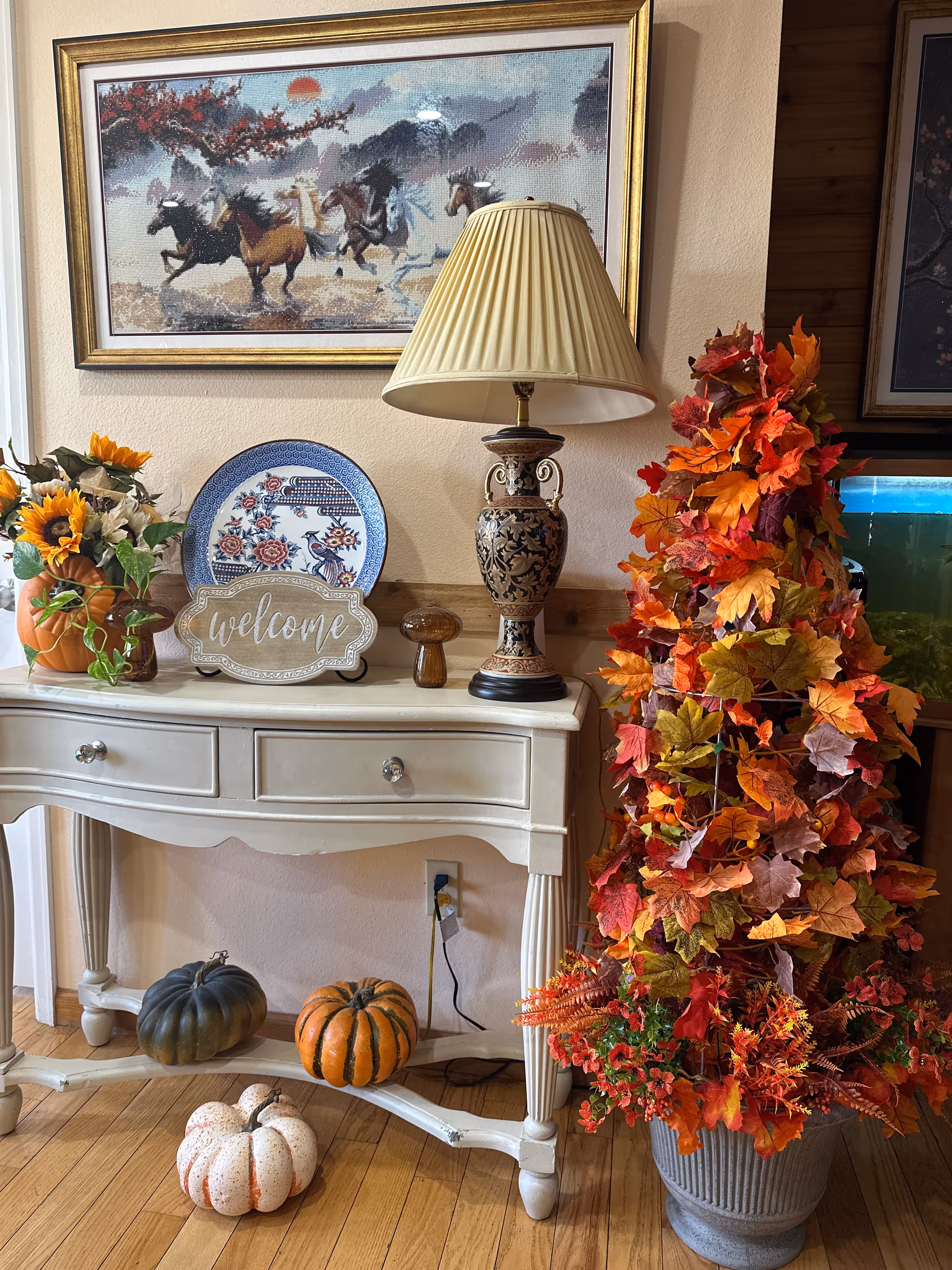 A decorative indoor scene featuring a white wooden console table with two drawers. On the table are a ceramic lamp with a beige pleated shade, a blue and white decorative plate, a small wooden mushroom figurine, a 'welcome' sign, and a vase with sunflowers and other flowers. Below the table are three small pumpkins in green, orange, and white. To the right of the table is a large potted arrangement of autumn leaves in shades of red, orange, and yellow. Above the table hangs a framed painting of running horses with a sunset background.