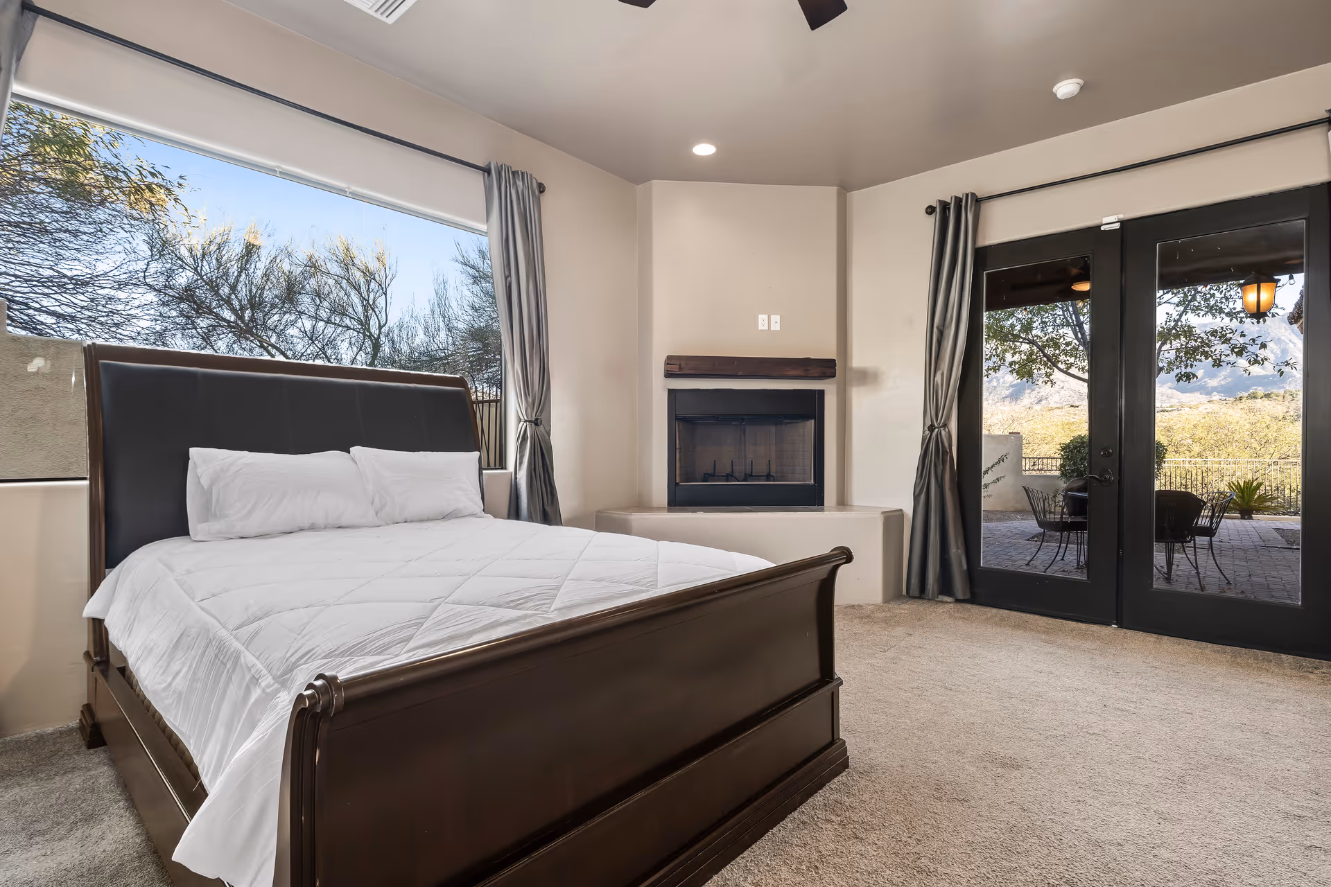 A bedroom with a large wooden bed featuring a white comforter and pillows. The room has beige walls and carpet, a corner fireplace, large windows with gray curtains, and glass double doors leading to an outdoor patio with chairs and a table. Trees and a scenic view are visible outside.