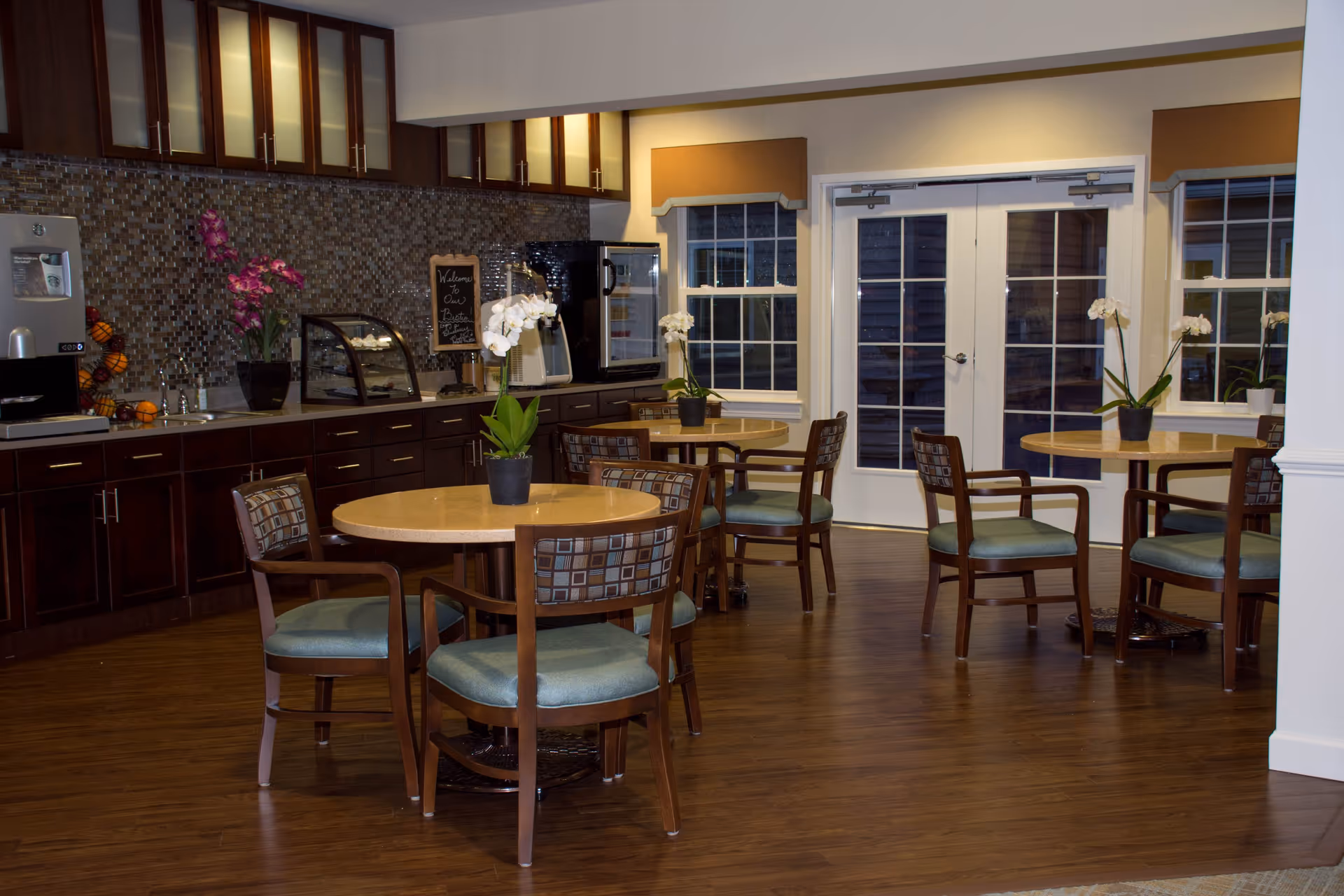 A cozy dining area with round wooden tables and cushioned chairs arranged neatly. Each table has a small potted orchid plant as decoration. The room features dark wood cabinetry with a mosaic tile backsplash, a coffee machine, and a display case on the counter. Large windows and glass doors allow natural light to enter the space.