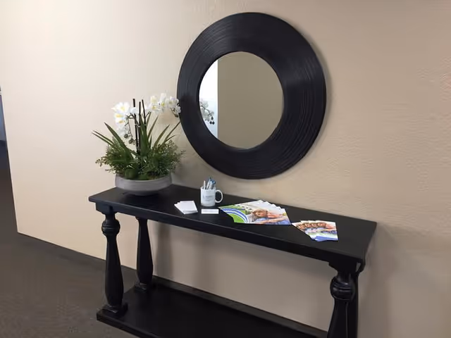 A black console table against a beige wall with a round black-framed mirror above it. On the table, there is a potted plant with white flowers, a white mug holding pens, some business cards, and brochures.